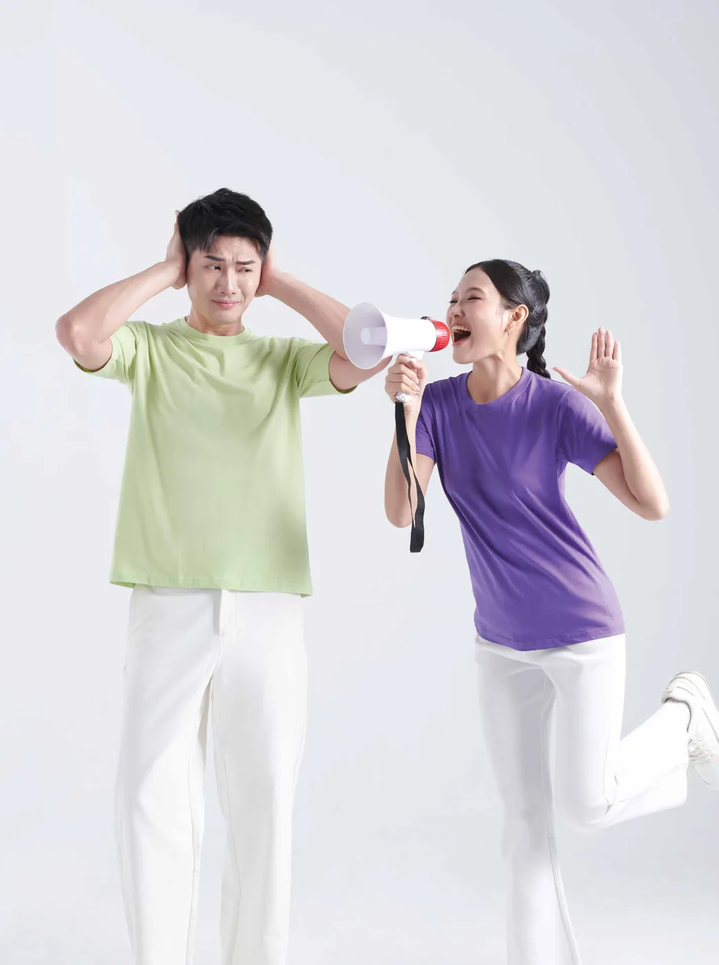 A man in a light green tee covers his ears while a woman in a purple tee shouts at him through a megaphone, against a white backdrop.