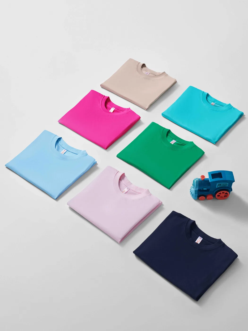 Folded kids' tees in beige, magenta, turquoise, light blue, green, light pink and navy laid out on a white surface beside a toy train.