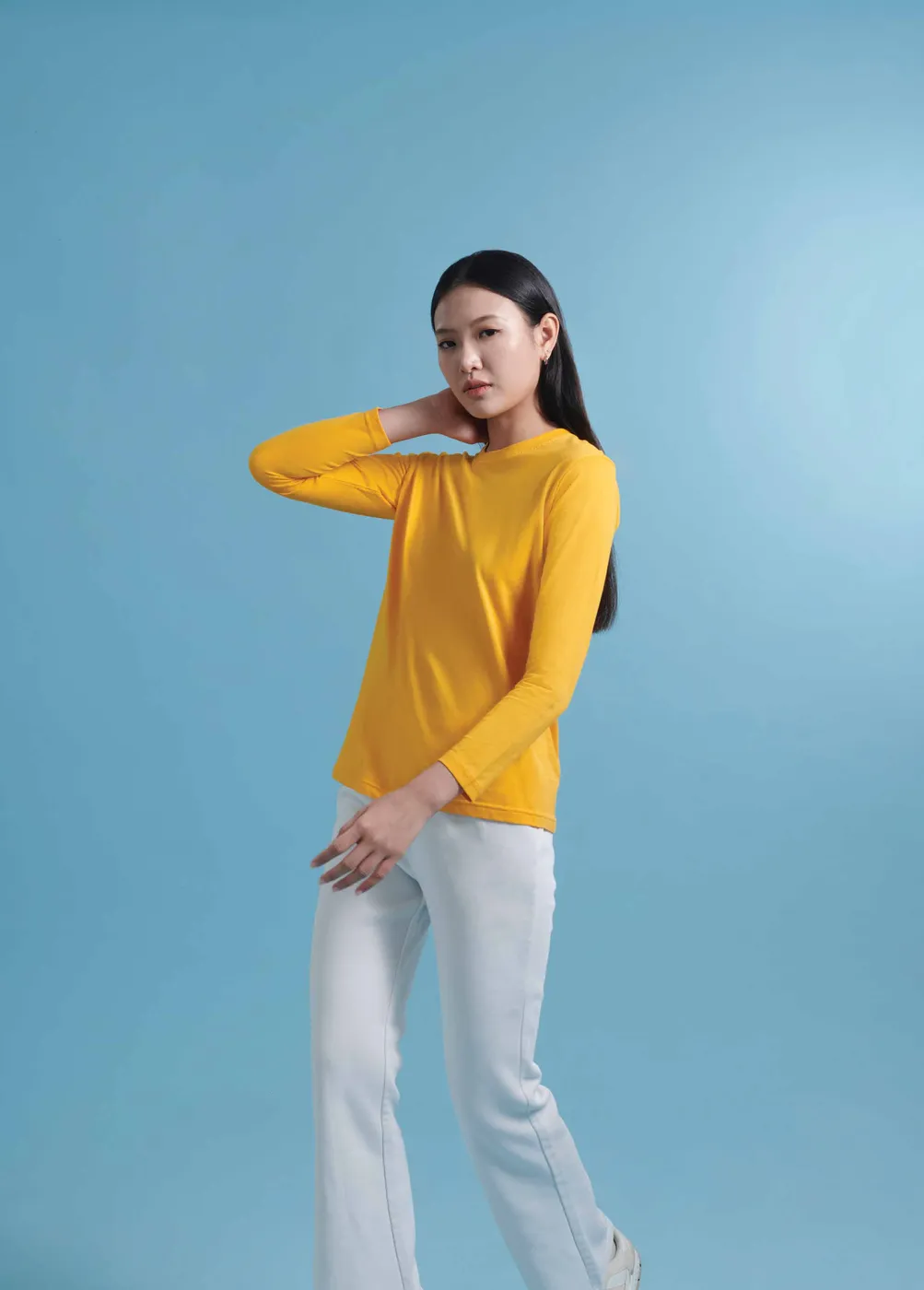 A woman in a yellow long sleeve tee and white jeans poses with one hand behind her head against a light blue backdrop.