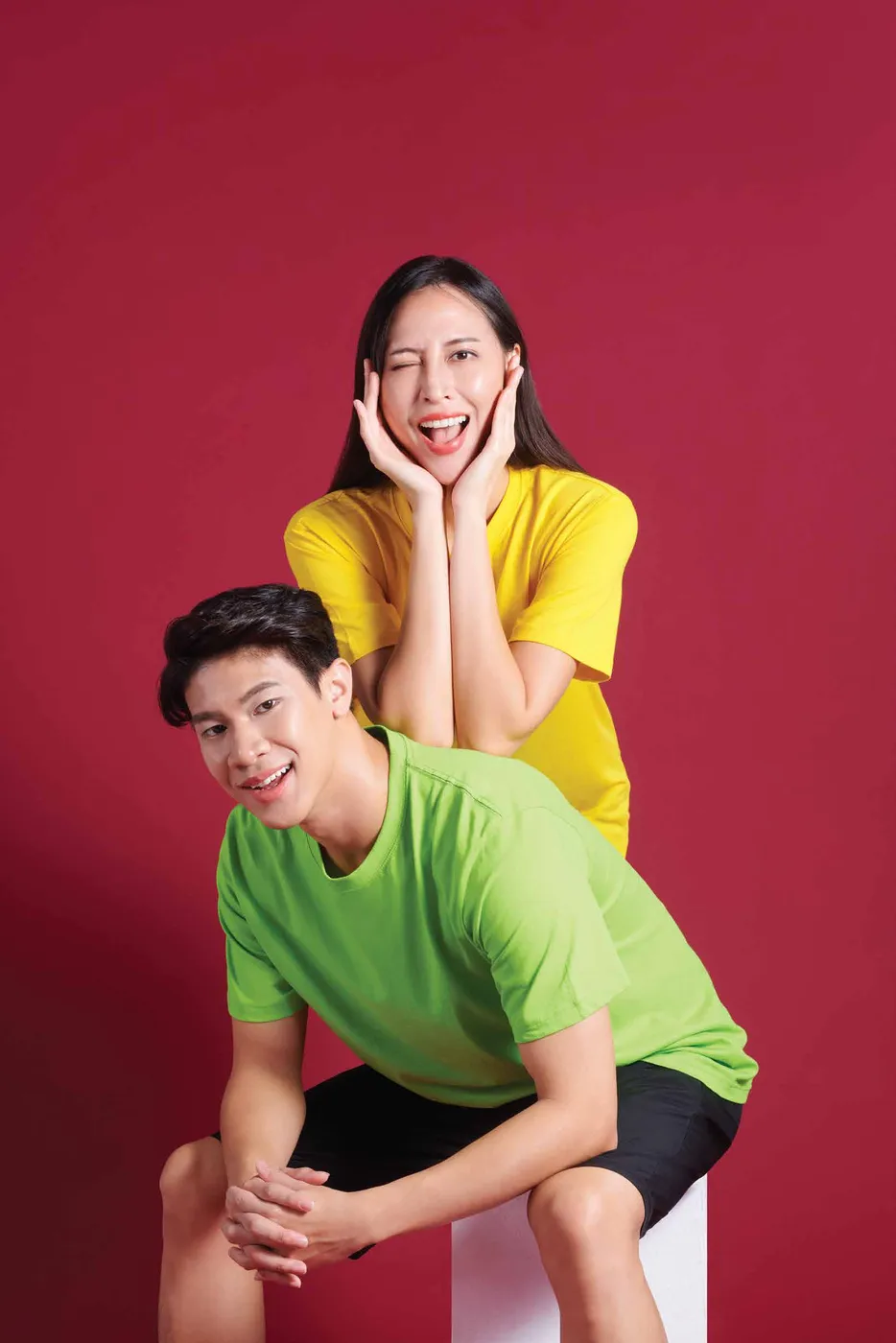 A man in a lime green tee crouches in front of a smiling woman in a yellow tee against a red backdrop.
