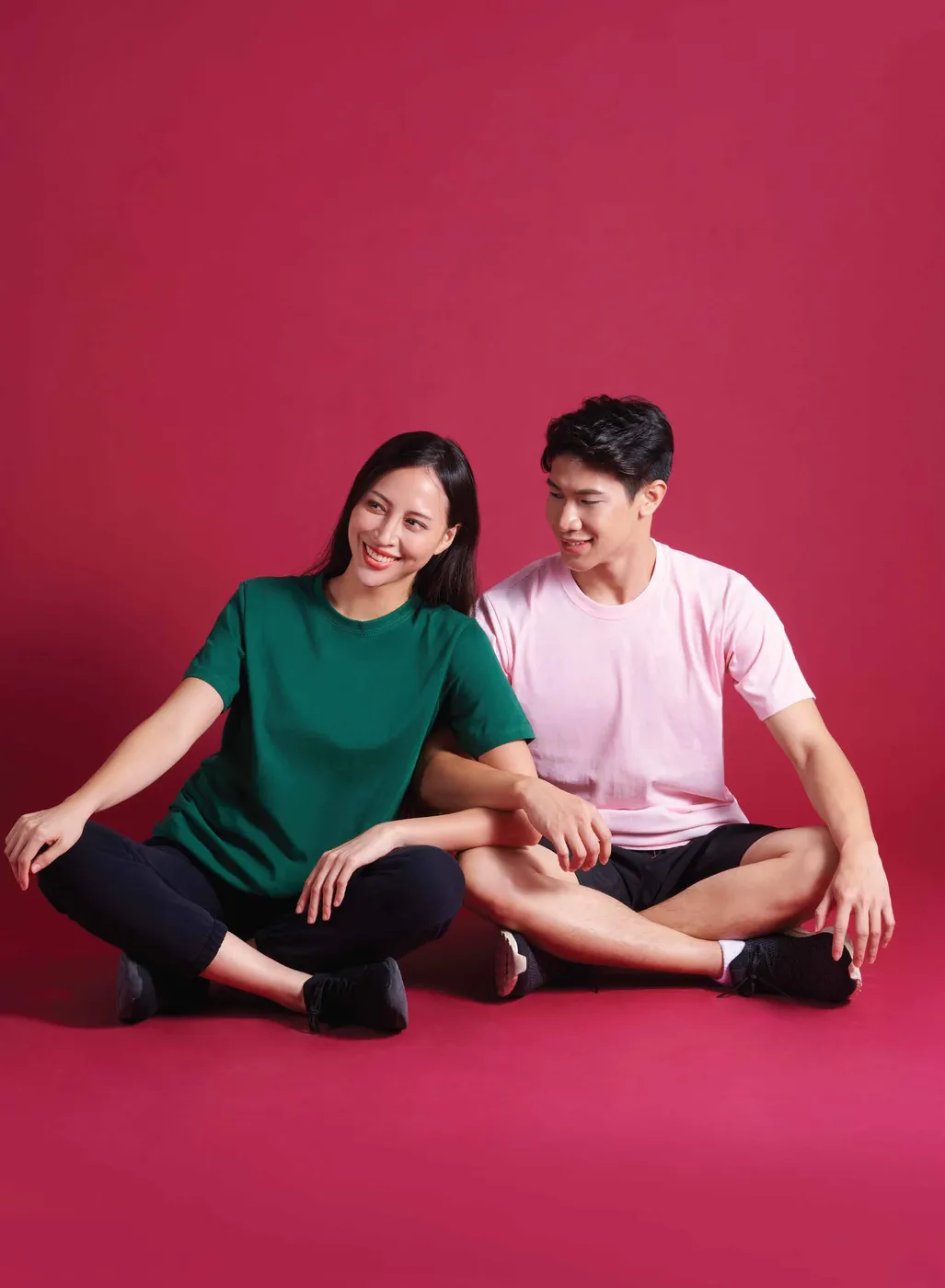 A woman in a forest green tee and a man in a light pink tee sit cross-legged on a red studio floor.